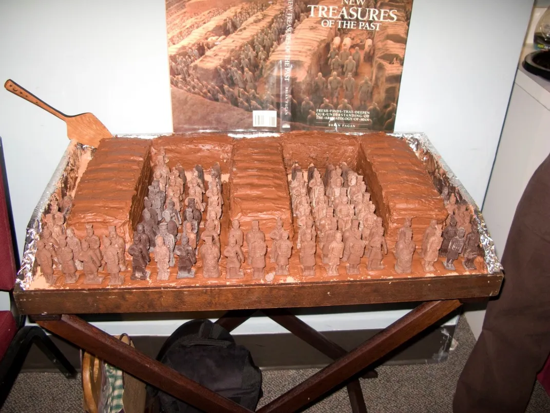 Brown, chocolate cake recreation of a Chinese tomb with brown, solid chocolate recreations of the terracotta warriors inside. The cake is on display on a brown, wood tray table. 